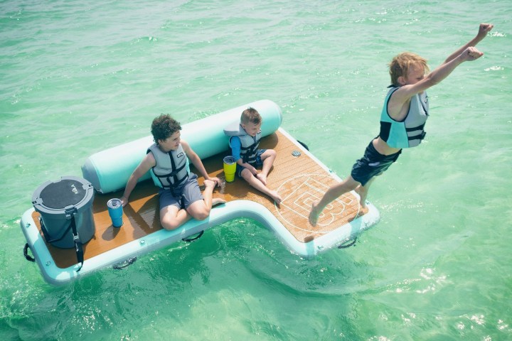 Three kids on a floating platform; one jumps into clear water.