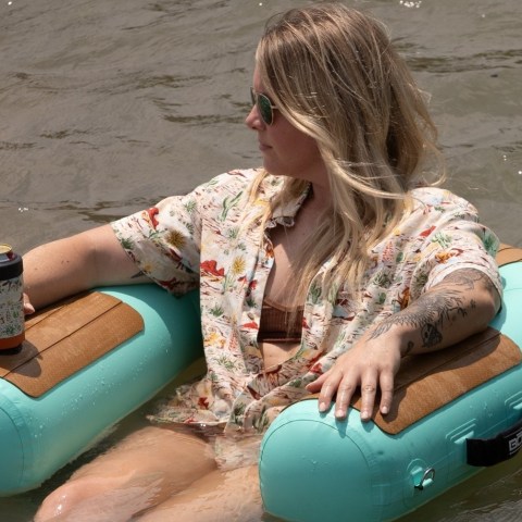 Person relaxing in a pool float with a drink, wearing sunglasses and a floral shirt.