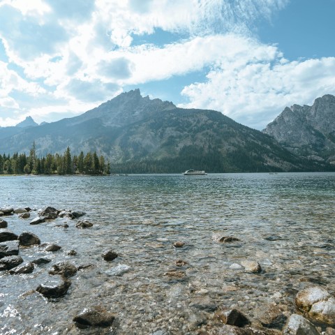 Rocky lakeshore with mountains and trees under a partly cloudy sky.