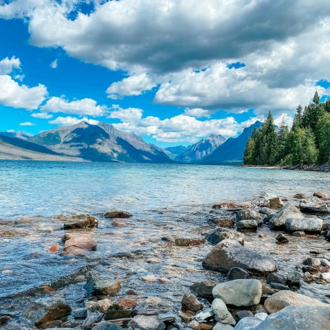 Rocky shoreline of a clear lake with mountains and cloudy sky.