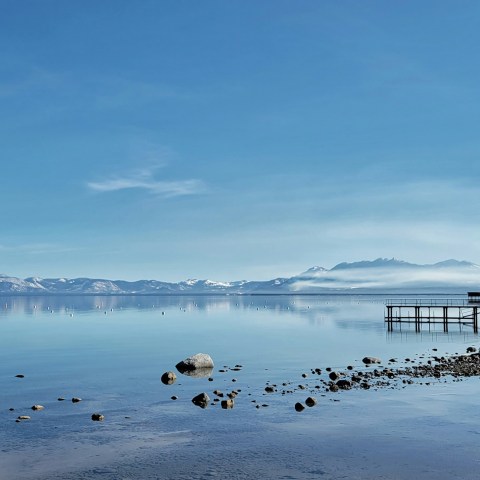Calm lake with distant mountains, a pier, and rocky shoreline under a clear blue sky.