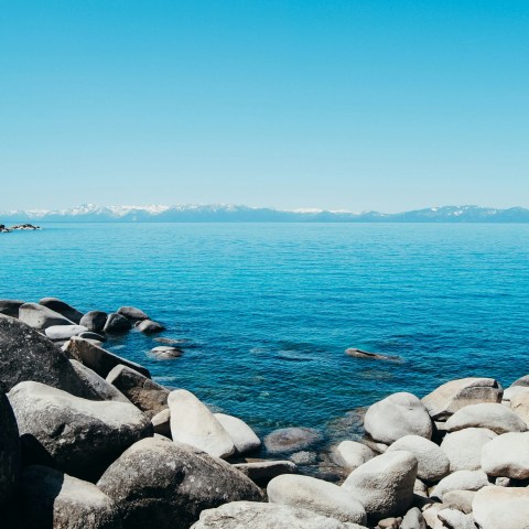 Rocky shoreline with calm blue lake and distant mountains under a clear sky.