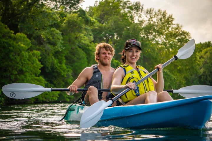 Two people kayaking on a calm river surrounded by greenery.